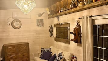 A cozy room with a wooden ceiling, a chandelier, a sofa with patterned cushions, a small table, and a window with curtains.