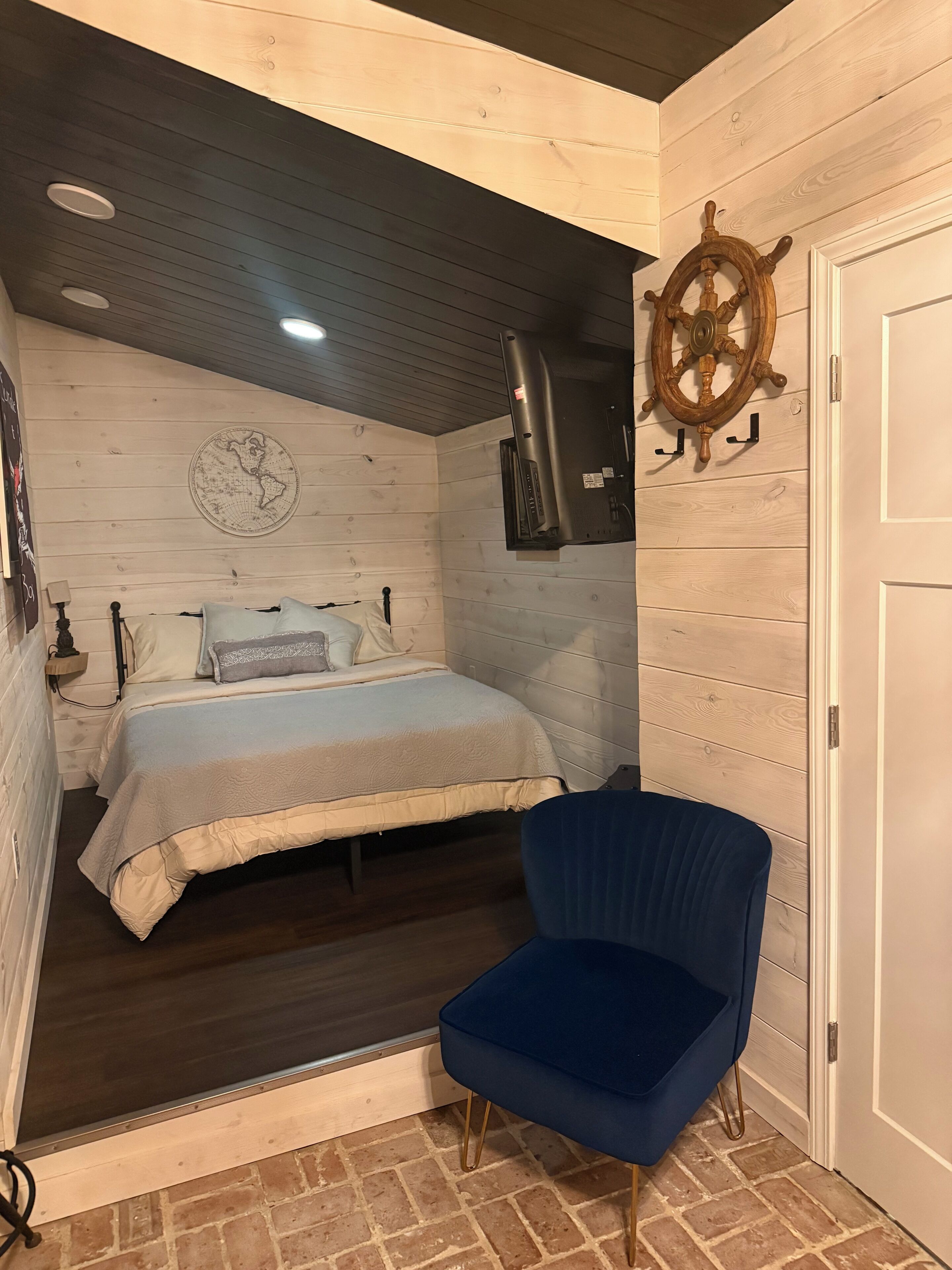 A bedroom with a bed, a chair, a map on the wall, and a ship's wheel.