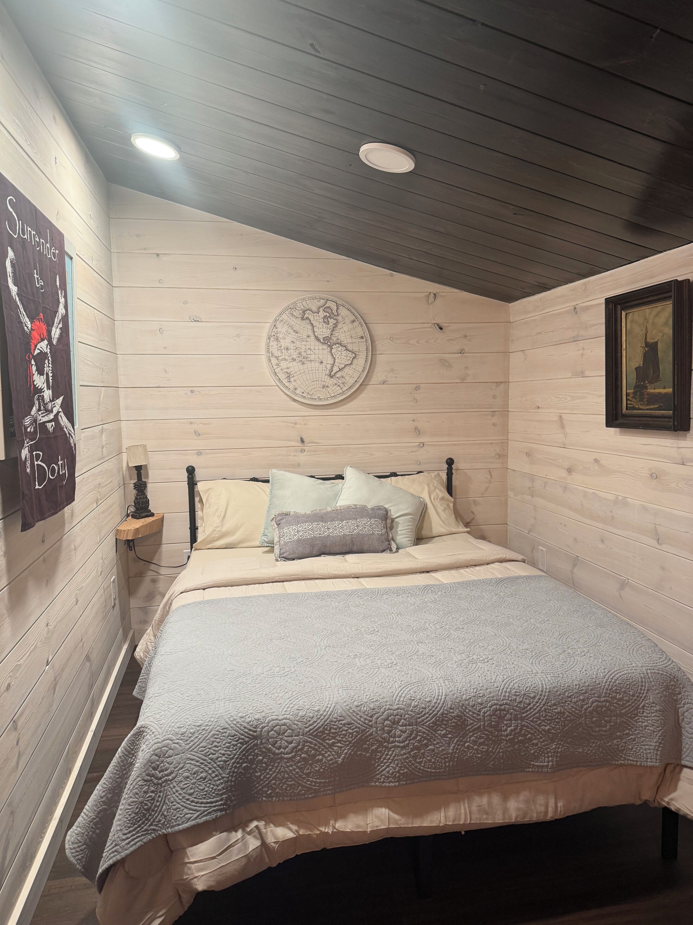 A bedroom with a wooden ceiling, a round wall map, a bed with a patterned blanket, and a framed picture on the wall.