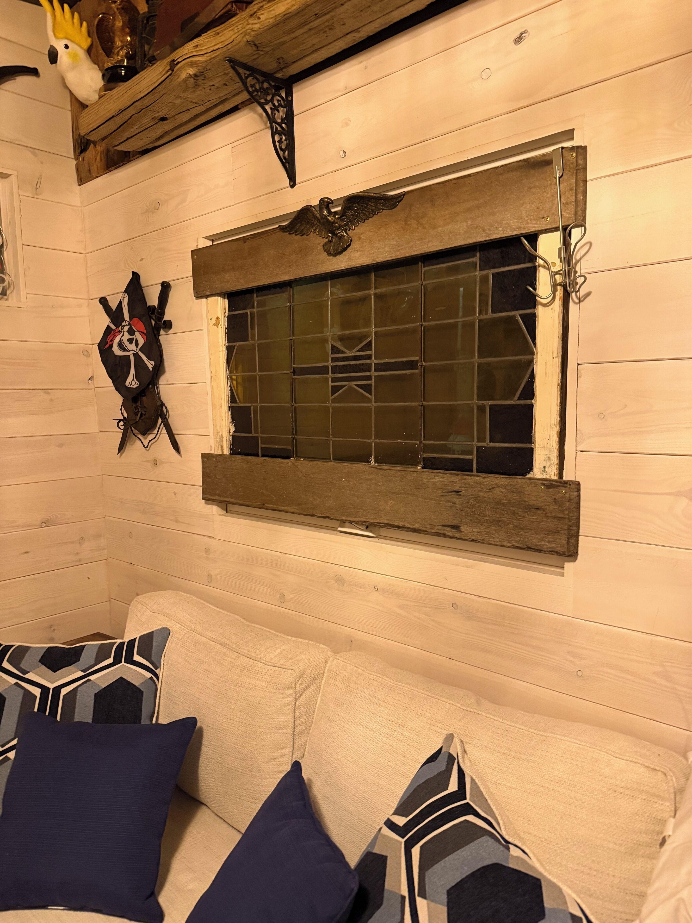 A cabin interior with a wooden wall, a window with a metal frame, a decorative skull and crossbones, and a couch with patterned pillows.