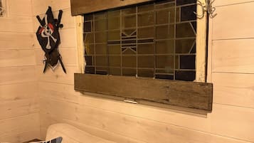 A cabin interior with a wooden wall, a window with a metal frame, a decorative skull and crossbones, and a couch with patterned pillows.