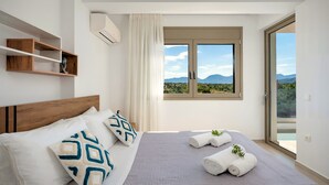 A bedroom with a bed, pillows, a window with a view, a shelf, and an air conditioning unit. - Matis Villa Private Pool (Vathi)