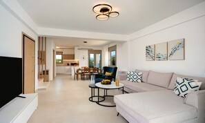 A modern living room with a sofa, a coffee table, and a TV. There is a dining area with a table and chairs. The room has a ceiling light fixture. - Matis Villa Private Pool (Vathi)