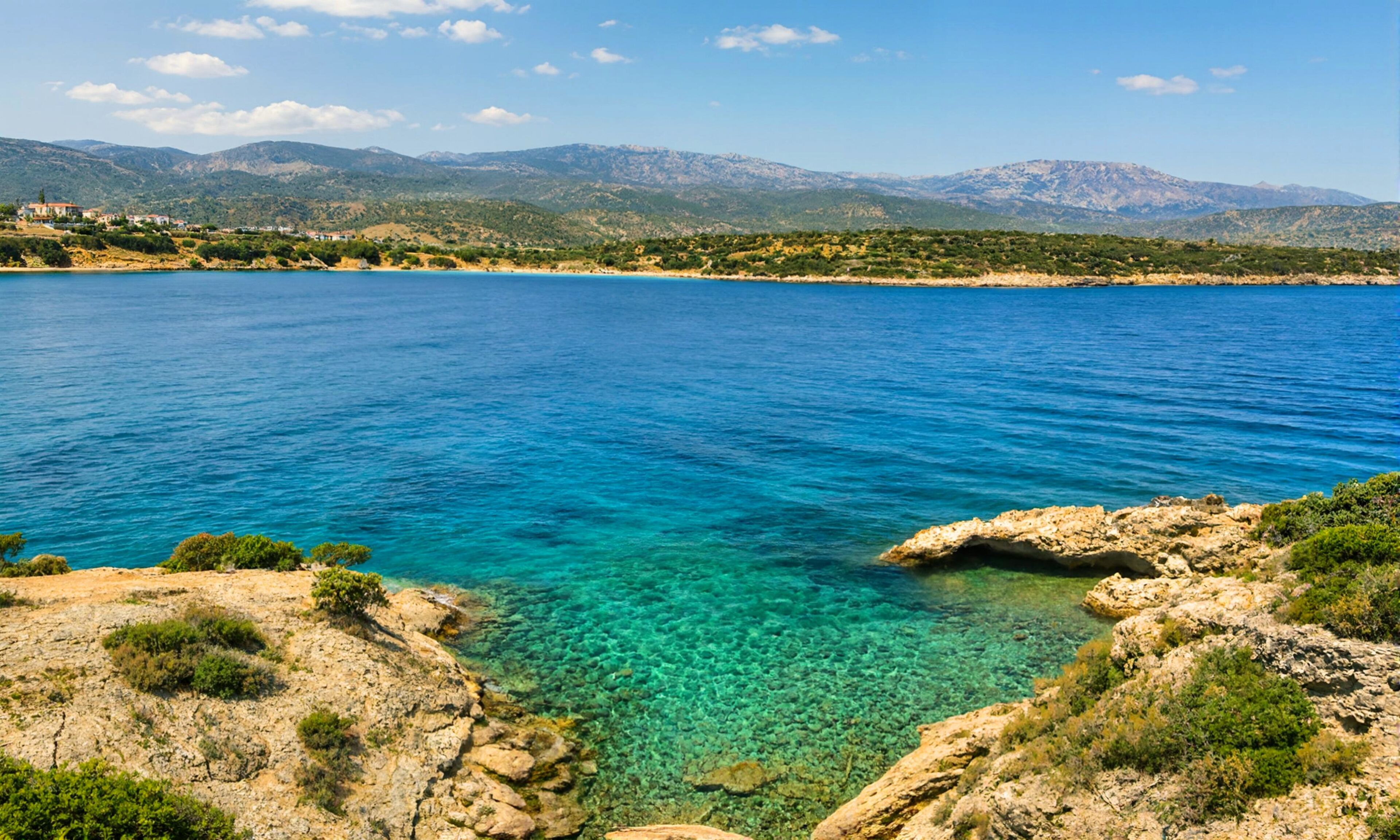 Un paysage côtier avec une mer bleue et limpide, des rivages rocheux et des montagnes à l’horizon.