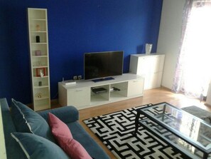 Ein Wohnzimmer mit einer Couch, einem Fernseher auf einem Schrank, einem Bücherregal und einem gemusterten Teppich.