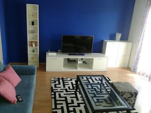 Ein Wohnzimmer mit blauem Sofa, einer weißen Unterhaltungseinheit mit Flachbildfernseher, einem gemusterten Teppich und einem Schrank.