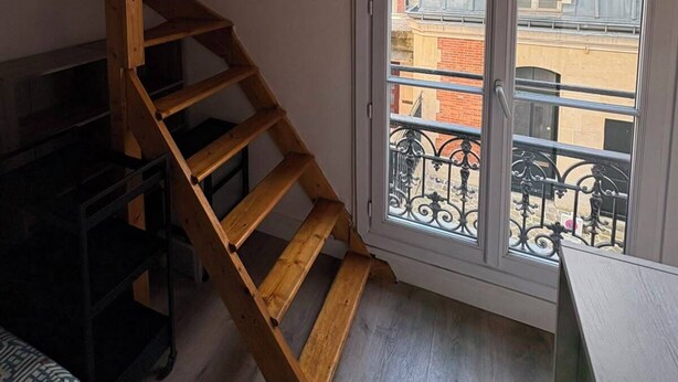 Ein Zimmer mit einer Holztreppe, einem Fenster mit Blick auf Gebäude und einem schwarzen Möbelstück.