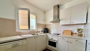 Eine Küche mit weißen Schränken, einem Geschirrspüler, einem Backofen, einem Fenster mit Jalousien und einer Pflanze auf der Arbeitsfläche.