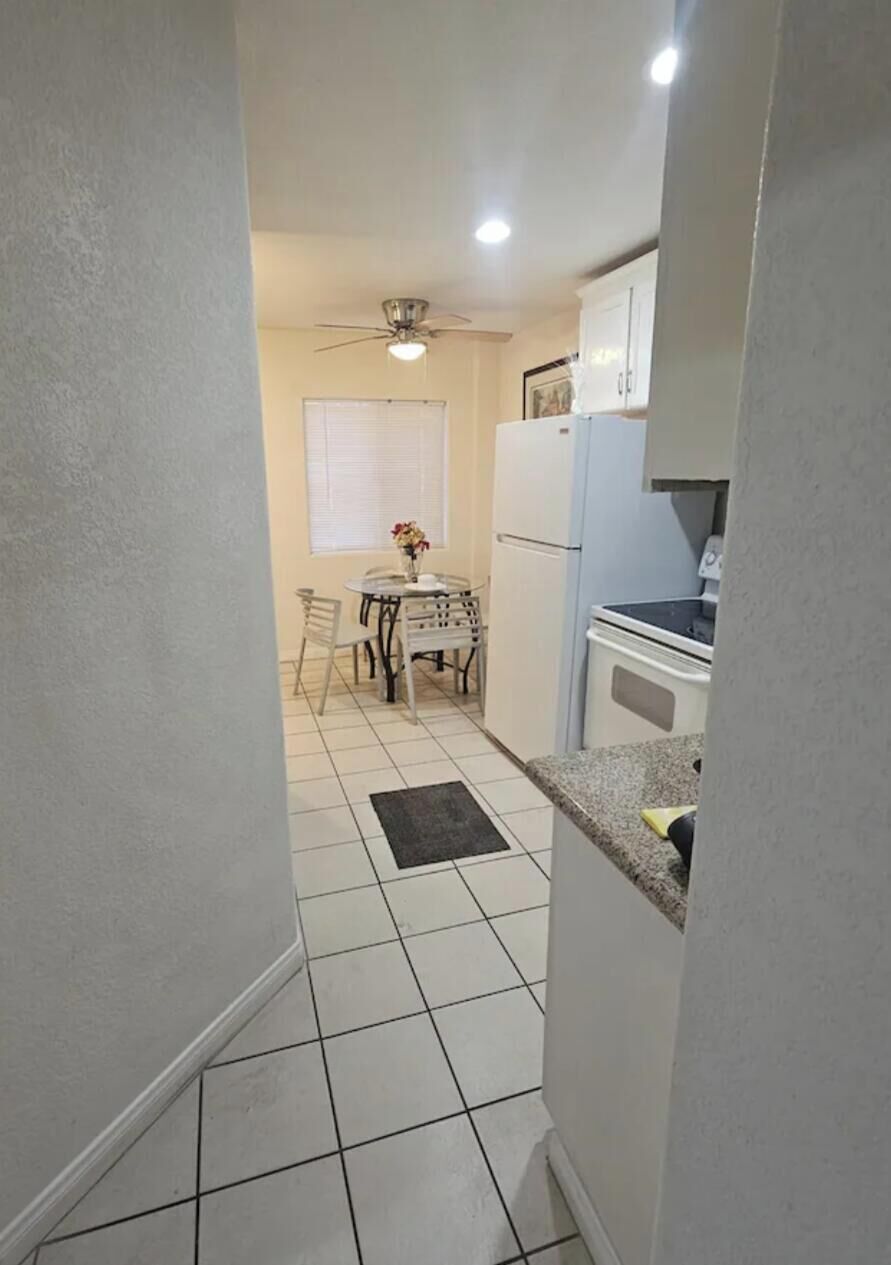 A narrow kitchen with white cabinets, a refrigerator, and a dining table with chairs.