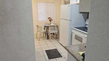 A narrow kitchen with white cabinets, a refrigerator, and a dining table with chairs.