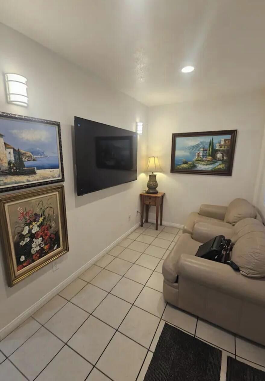 A living room with a beige sofa, a flat-screen TV mounted on the wall, a lamp on a side table, and framed artwork on the walls.