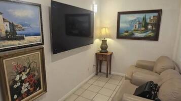 A living room with a beige sofa, a flat-screen TV mounted on the wall, a lamp on a side table, and framed artwork on the walls.