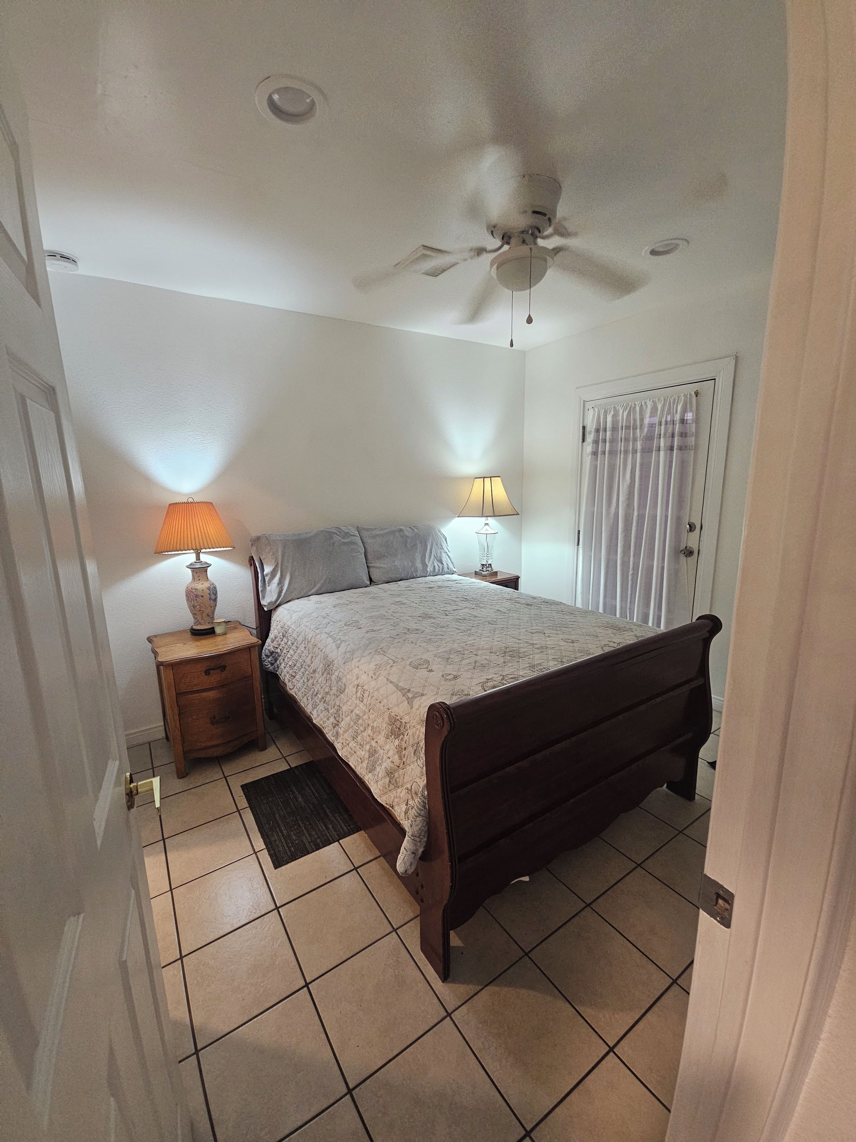 A bedroom with a bed, two bedside lamps, a ceiling fan, a door with a curtain, and a tiled floor.