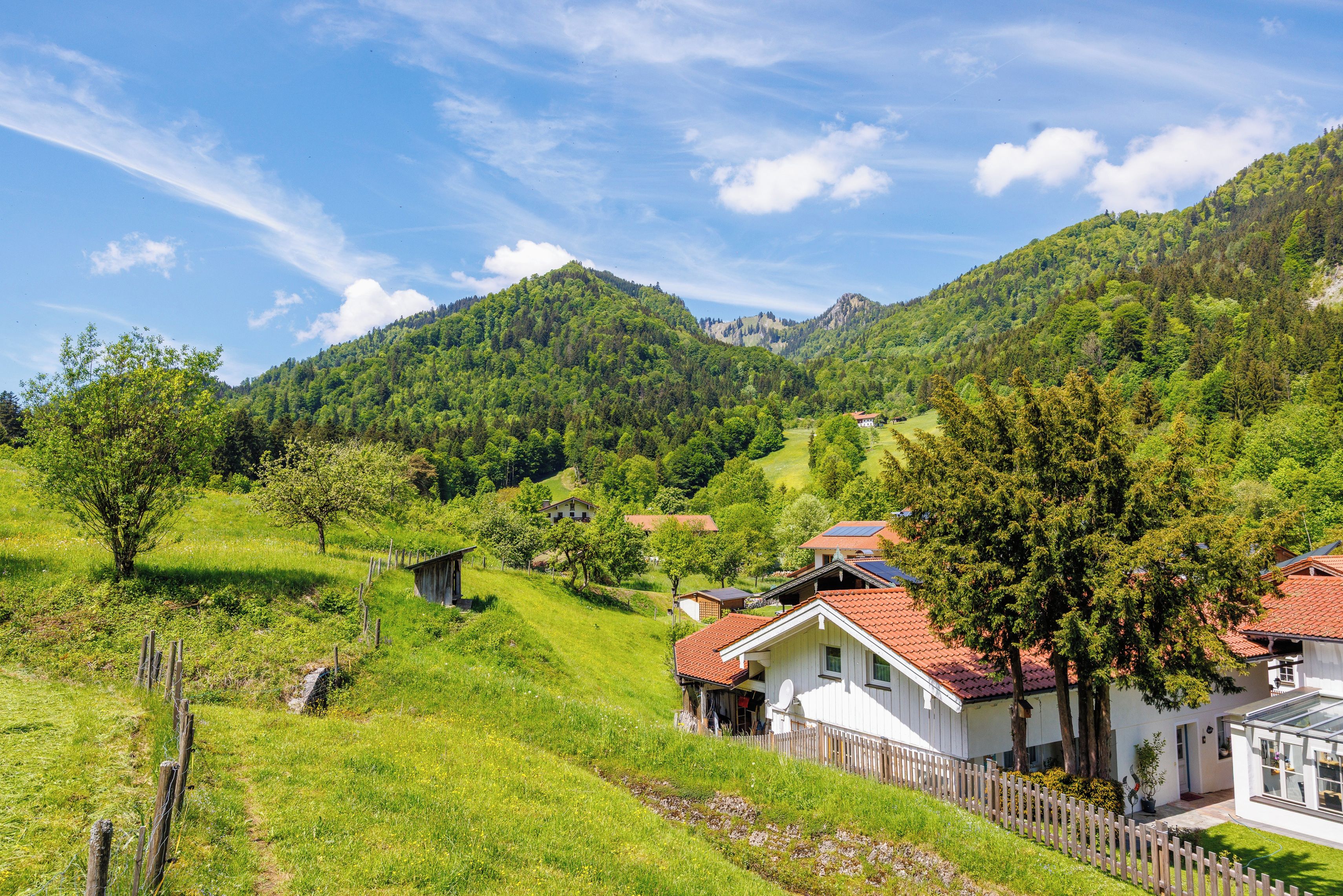 A rural landscape with a white house featuring a red roof, surrounded by lush greenery and hills.