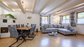 A modern living room with a wooden dining table, grey upholstered chairs, a grey sofa, and large windows with curtains. - Hinterseer Vacation Home (Ruhpolding)