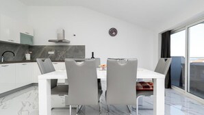 Et moderne køkken med hvide skabe, en marmor bordplade og et spiseområde med et hvidt bord og grå stole.