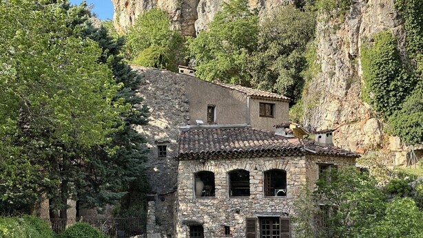 A stone building with a tiled roof, surrounded by greenery and cliffs.