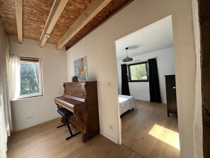 A room with a wooden floor, a piano, a bed with a patterned bedspread, a map on the wall, and a window with curtains. - Wooden house & serenity near Guingamp (Ploumagoar)