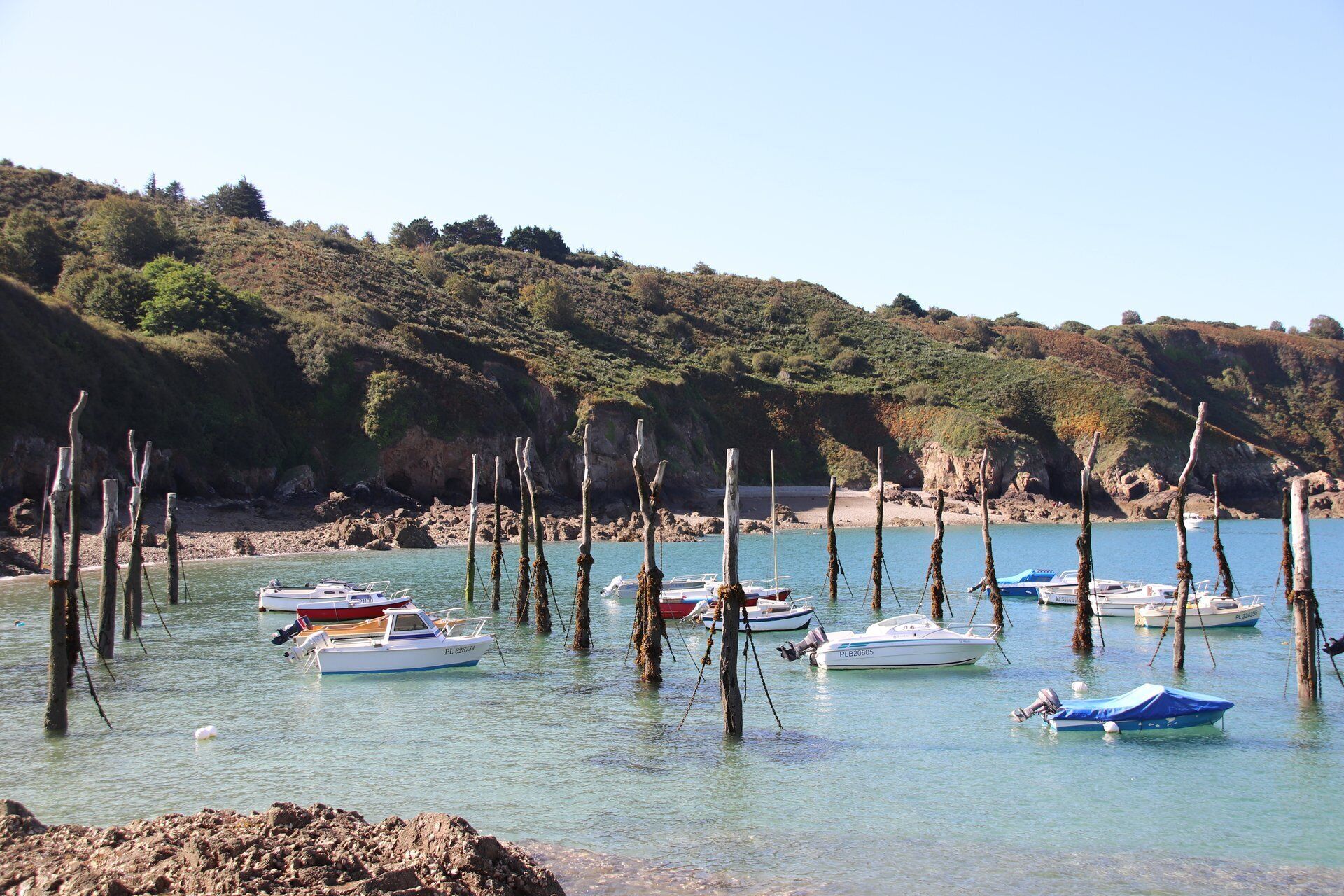 Une zone côtière où des bateaux sont amarrés près de piliers en bois, l’eau est claire et le rivage rocheux est bordé de végétation.