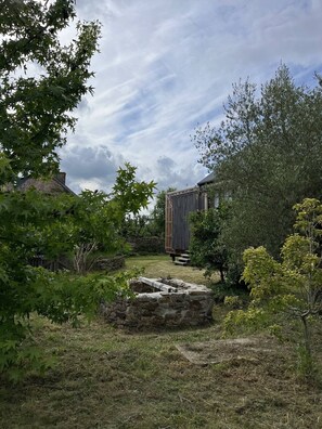 A rustic stone well, a wooden shed, and a brick building in a garden with trees and grass. - Wooden house & serenity near Guingamp (Ploumagoar)