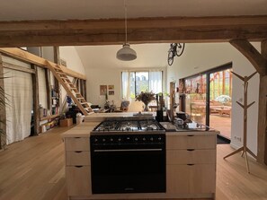 Una cocina con estufa a gas, vigas de madera, zona de comedor con mesa y sillas, y vistas a través de un ventanal.