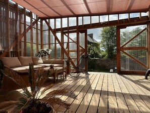 A spacious wooden deck with a cushioned seating area, large windows, and a view of the surrounding greenery. - Wooden house & serenity near Guingamp (Ploumagoar)