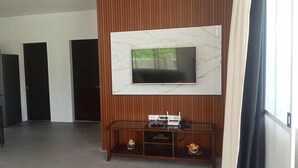 Une chambre d’hôtel moderne dotée d’un grand téléviseur à écran plat fixé sur un mur en marbre, d’un meuble de rangement en bois en dessous et d’un coin repas avec une table et des chaises.