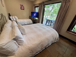 A double bed with white bedding, a television on a stand, and a view of an outdoor patio with a table and chairs. - Barbet Room - Staynest Guesthouse (Chartwell)