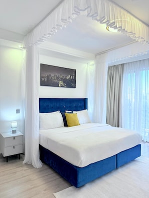 A bedroom with a bed, a nightstand, a lamp, a cityscape painting, and sheer curtains. - 108 riverside (Nairobi)