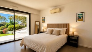 A modern bedroom with a large bed, two bedside tables with lamps, an air conditioning unit, and a view of a garden through a sliding door. - Contemporary villa with ocean views and a pool, sleeps 10, in Cavalaire-sur-Mer (Cavalaire-sur-Mer)