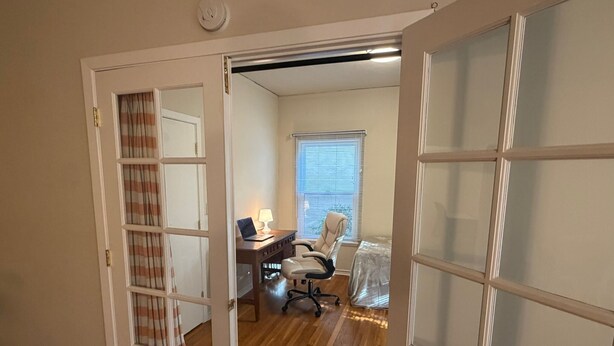 Ein Zimmer mit Holzboden, Schreibtisch mit Lampe, Stuhl und einem Fenster mit Jalousien.