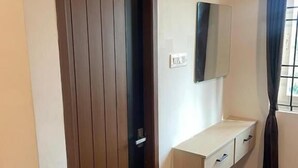 Una habitación con puerta de madera, un armario pequeño blanco, un espejo y una ventana con cortinas.