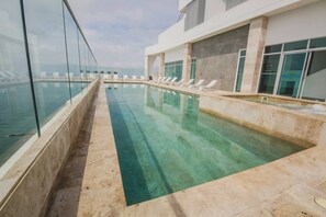 Ein Schwimmbecken mit klarem Wasser, umgeben von Liegestühlen und einem modernen Gebäude mit großen Fenstern.