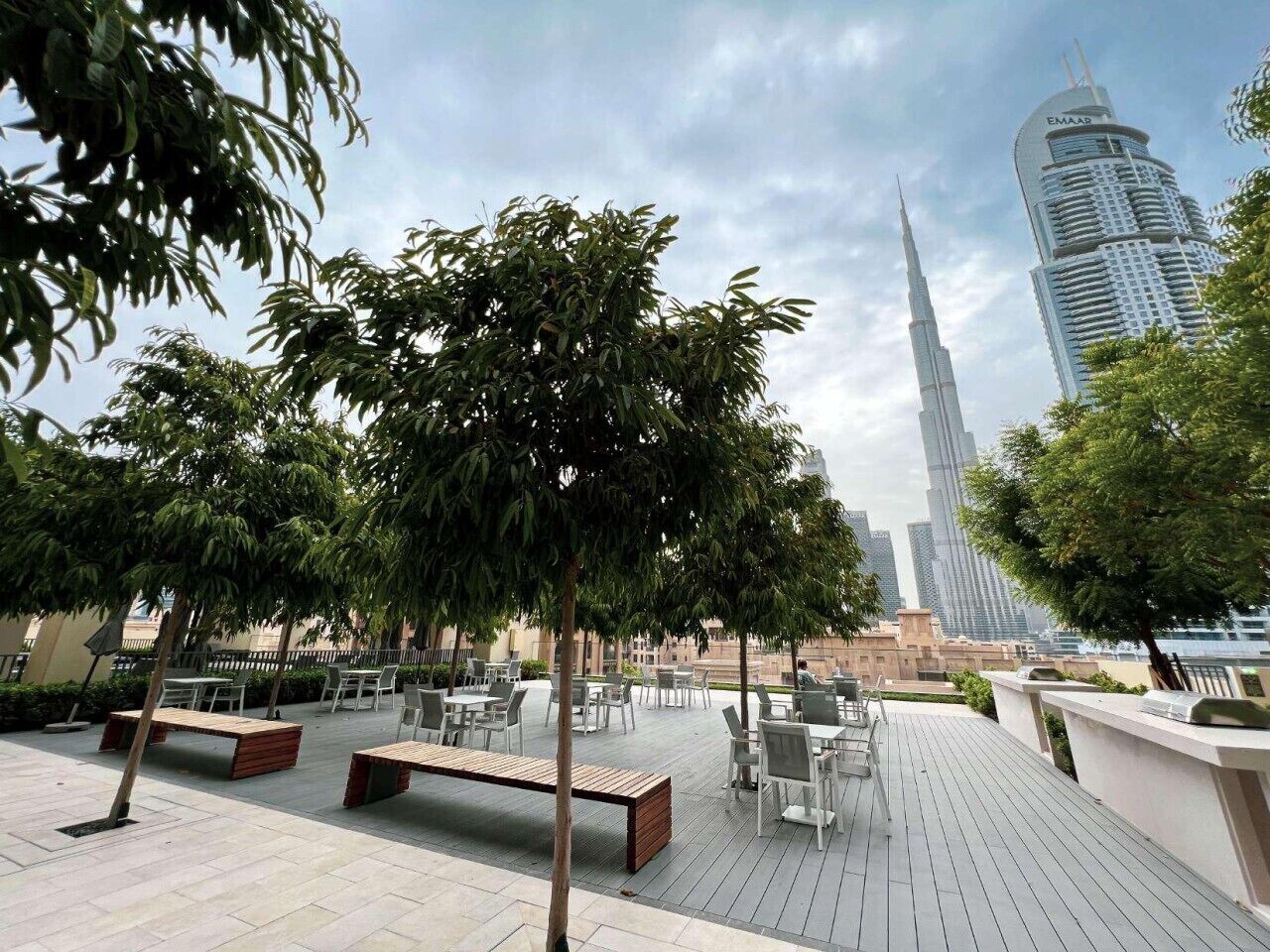 Un espace extérieur aménagé avec des tables et des chaises, entouré d’arbres, et offrant une vue sur un grand immeuble en arrière-plan.