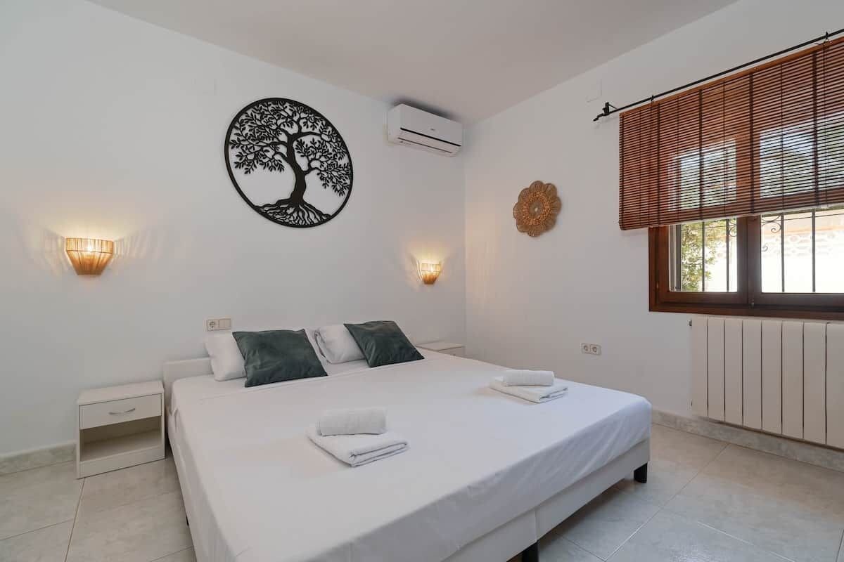 A bedroom with a bed, two wall sconces, an air conditioner, a window with blinds, and a decorative wall piece.
