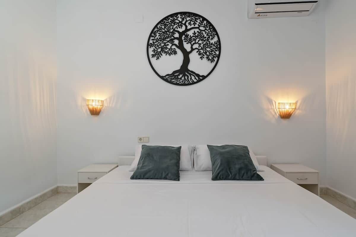 Une chambre à coucher comprenant un lit, deux tables de chevet, deux lampes fixées au mur et une œuvre d’art représentant un arbre.
