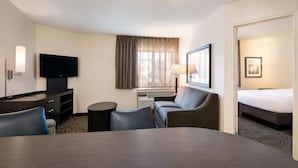 A hotel room with a flat-screen TV, a sofa, a bed, a dining table, and a window with curtains.