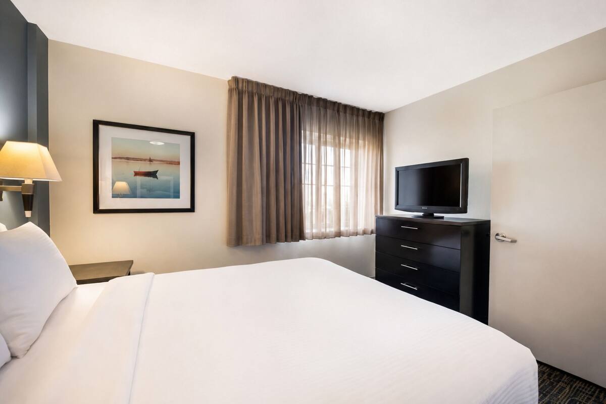 Une chambre d’hôtel avec un lit, un téléviseur posé sur une commode, un tableau encadré au mur et une fenêtre avec des rideaux.