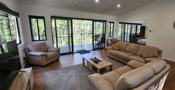 A spacious living room with a sofa, armchair, and a coffee table. Large windows provide a view of the outdoors.
