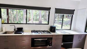 Uma cozinha moderna com fogão a gás, forno e pia, grandes janelas com vista para uma floresta e uma geladeira branca.