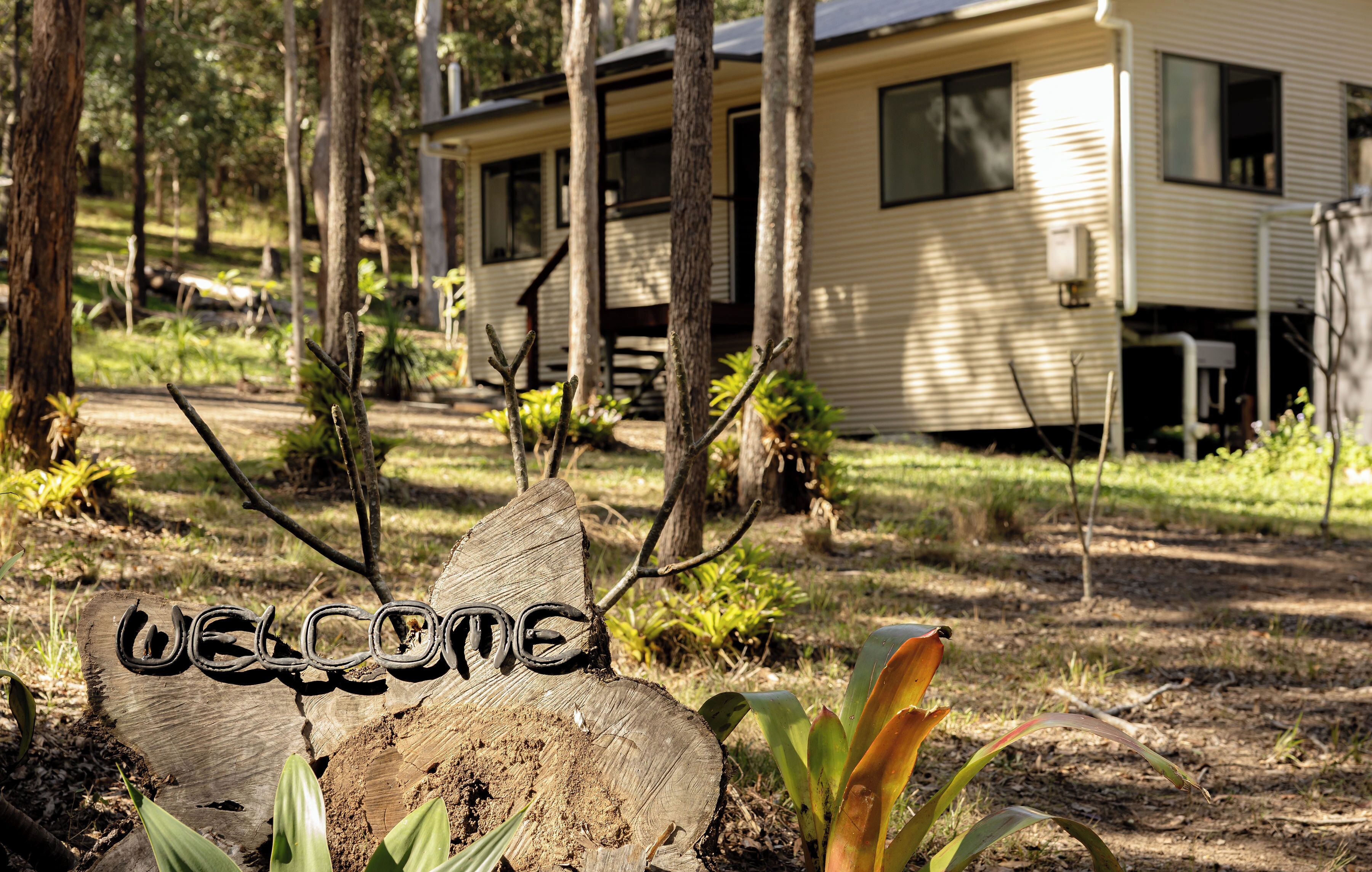 A wooden sign with 'WELCOME' carved into it, surrounded by plants and trees, with a mobile home in the background.