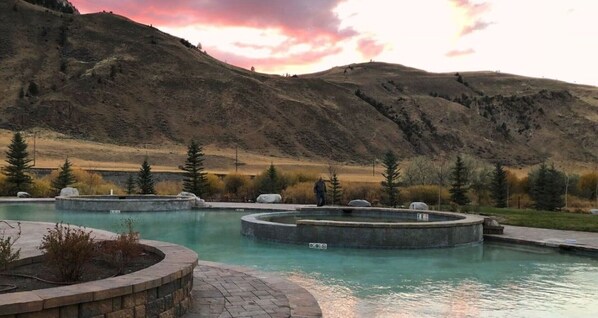 Ein Pool mit kreisförmigem Design, umgeben von einer begrünten Anlage mit Bäumen und einem Bergpanorama im Hintergrund.
