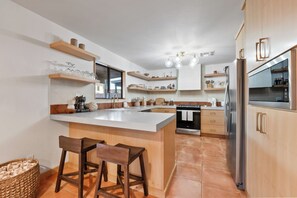 A modern kitchen with a central island, bar stools, wooden shelves, and a stainless steel refrigerator. - Desert Sonoran Retreat, 4-bedroom house with AC, "Heated pool", EV charger (Green Valley)