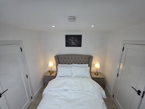 A bedroom with a tufted headboard, white bedding, two bedside lamps, a framed artwork above the bed, and white doors on either side.