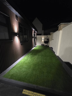 A narrow pathway with artificial grass, a wall-mounted light fixture, and a door leading to a building with vertical siding.