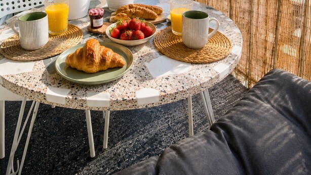 En balkong med ett bord dukat för frukost, med bland annat croissanter, jordgubbar och juice, och utsikt över staden.