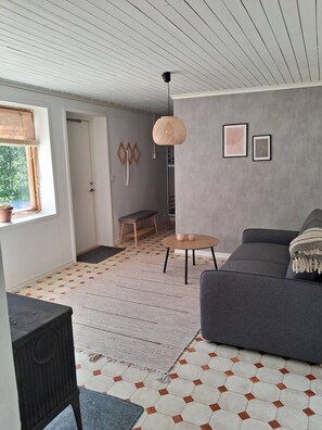 Uma sala de estar moderna com um sofá cinzento, uma mesa de madeira redonda, um candeeiro pendente trançado e um tapete estampado.