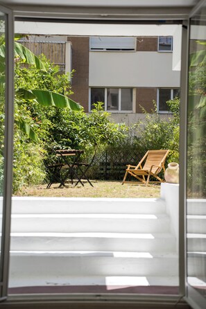 A view through a window of a backyard with a wooden chair, a small table, and various plants. - Charming Parisian Apartment with Garden View (Paris)