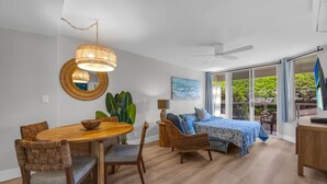 A modern living room with a dining table, wicker chairs, a large plant, a ceiling fan, and a bed with a view. - 2 2 New Designer Beachside Resort Vistas (Kihei)