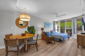 A modern living room with a dining table, wicker chairs, a large plant, a ceiling fan, and a bed with a view. - 2 2 New Designer Beachside Resort Vistas (Kihei)